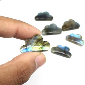 Peut inclure: Plusieurs pierres précieuses en labradorite en forme de nuage. L'une est tenue entre les doigts, révélant des reflets bleus, verts et jaunes. Les autres pierres sont dispersées, chacune avec des variations de couleurs uniques.