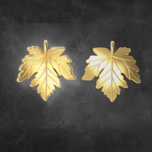 May include: Two gold-colored maple leaf pendants. The pendants are detailed with veining and a loop for hanging. The leaves are set against a dark gray background. The pendants are likely for jewelry making or as decorative charms.