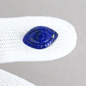 May include: A dark blue, eye-shaped gemstone with detailed carvings. The gemstone has a central eye design and surrounding patterns. The stone is held against a white background.