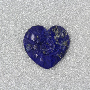 Puede incluir: Una piedra de lapisl&aacute;zuli azul oscuro en forma de coraz&oacute;n, con detalles tallados. La piedra presenta un dise&ntilde;o de ojo en el centro, rodeado de l&iacute;neas radiales. La piedra est&aacute; sobre un fondo plateado.