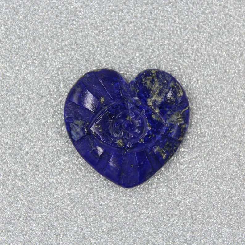 Puede incluir: Una piedra de lapisl&aacute;zuli azul oscuro en forma de coraz&oacute;n, con detalles tallados. La piedra presenta un dise&ntilde;o de ojo en el centro, rodeado de l&iacute;neas radiales. La piedra est&aacute; sobre un fondo plateado.