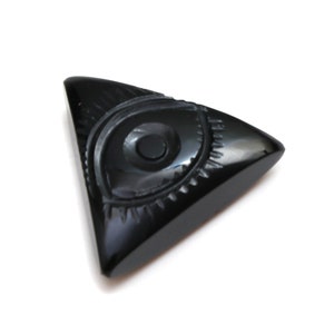 Puede incluir: Botón triangular negro brillante con diseño de ojo. El ojo está tallado con un iris detallado y elementos texturizados circundantes. El botón tiene una superficie lisa y pulida y una forma ligeramente redondeada.