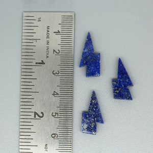 Pode incluir: Quatro peças de pedra preciosa de lápis-lazúli azul, cortadas em forma de raio, com manchas douradas. Uma régua de metal com marcações em centímetros está ao lado das pedras preciosas. A régua está marcada "MADE IN INDIA".