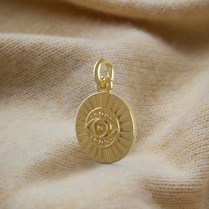 May include: A gold-coloured pendant featuring an eye design. The round pendant has a sunburst pattern around the eye. The eye is detailed with an iris, pupil, and eyelashes. It has a small loop at the top for a chain.