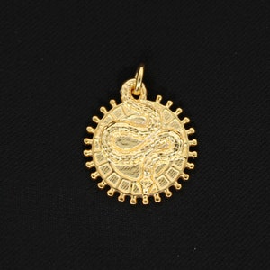 以下が含まれることがあります： 円形のフレームの中に、詳細なヘビのデザインが施されたゴールドカラーのペンダント。ペンダントはテクスチャ加工された表面を持ち、チェーンを通すための小さなループが上部にあります。円の外側の縁にはサンバーストのデザインがあります。