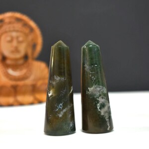 May include: Two green and brown moss agate obelisks with white veining. The obelisks are tapered and have a polished finish. The background includes a blurred wooden Buddha statue.