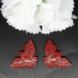 May include: Two carved, reddish-brown leaf-shaped pendants with detailed veining. The pendants are set against a black background, with a white flower in the upper portion of the image. The leaves are symmetrical and appear to be made of a hard material.
