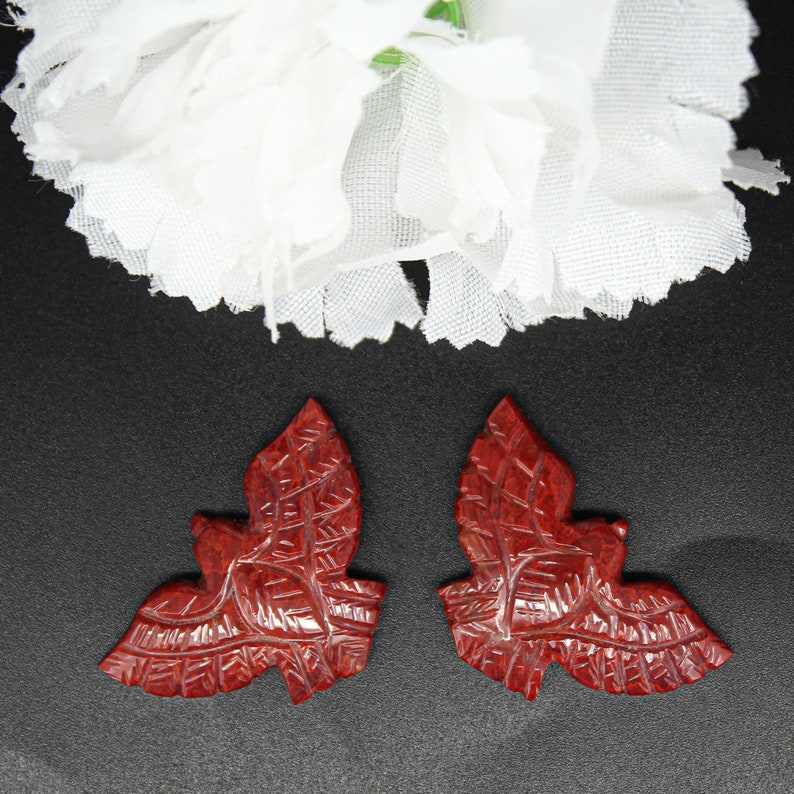 May include: Two carved, reddish-brown leaf-shaped pendants with detailed veining. The pendants are set against a black background, with a white flower in the upper portion of the image. The leaves are symmetrical and appear to be made of a hard material.