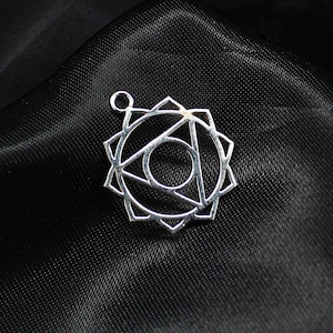 Może przedstawiać: Srebrny wisiorek o geometrycznym wzorze, z centralnym okręgiem, trójkątem i otaczającym wzorem w kształcie gwiazdy. Wisiorek ma małą pętlę na górze na łańcuszek. Wisiorek jest prezentowany na czarnej, teksturowanej tkaninie.