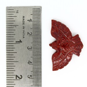 May include: A carved, reddish-brown stone bird figurine. The figurine is approximately 5 cm long. Intricate details are carved into the wings and body. The bird is positioned on a white surface next to a metal ruler.