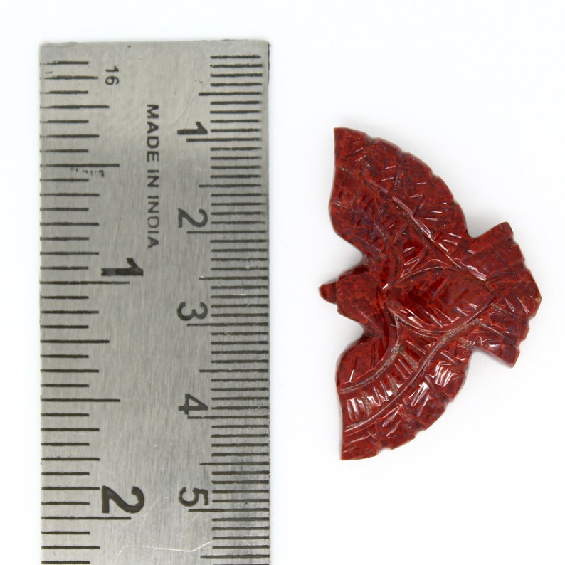 May include: A carved, reddish-brown stone bird figurine. The figurine is approximately 5 cm long. Intricate details are carved into the wings and body. The bird is positioned on a white surface next to a metal ruler.