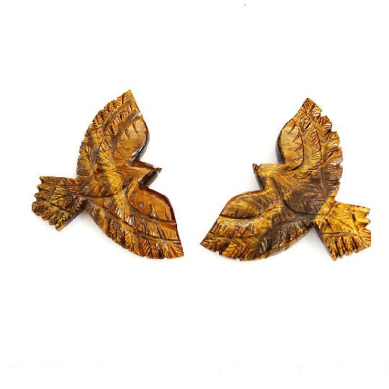 May include: Two wooden bird sculptures in flight, each with detailed wing and feather carvings. The birds are a warm brown color with darker brown and yellow accents, set against a white background.