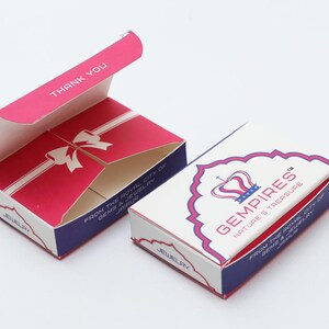 May include: Two jewellery boxes, one open and one closed, on a white surface. The open box is red with a white bow design and the words "THANK YOU". The closed box is white with the text "GEMPIRES NATURE'S TREASURE".