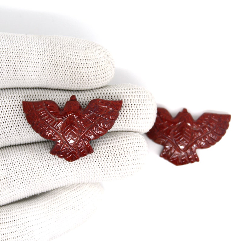 May include: Two carved red jasper eagle pendants with outstretched wings. The pendants have detailed patterns carved into the wings and body. The background is white.