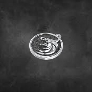 May include: A silver pendant with a wolf head emblem in a circular frame. The wolf head is a detailed, three-dimensional design, set against a textured, dark background. The pendant has a small loop for a chain.