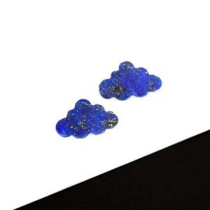 May include: Two cloud-shaped lapis lazuli gemstones. The gemstones are a deep blue colour with flecks of gold throughout. The clouds are small and have a rounded shape. They are set against a white background.