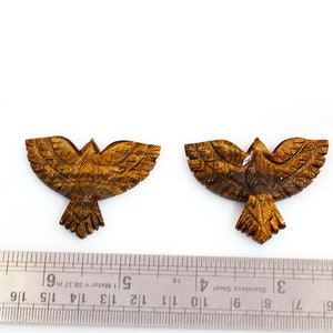 May include: Two carved wooden bird figurines with outstretched wings. The birds are a warm brown color with detailed feather carvings. A ruler is in the foreground, indicating the size of the birds.