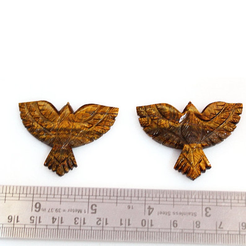 May include: Two carved wooden bird figurines with outstretched wings. The birds are a warm brown color with detailed feather carvings. A ruler is in the foreground, indicating the size of the birds.