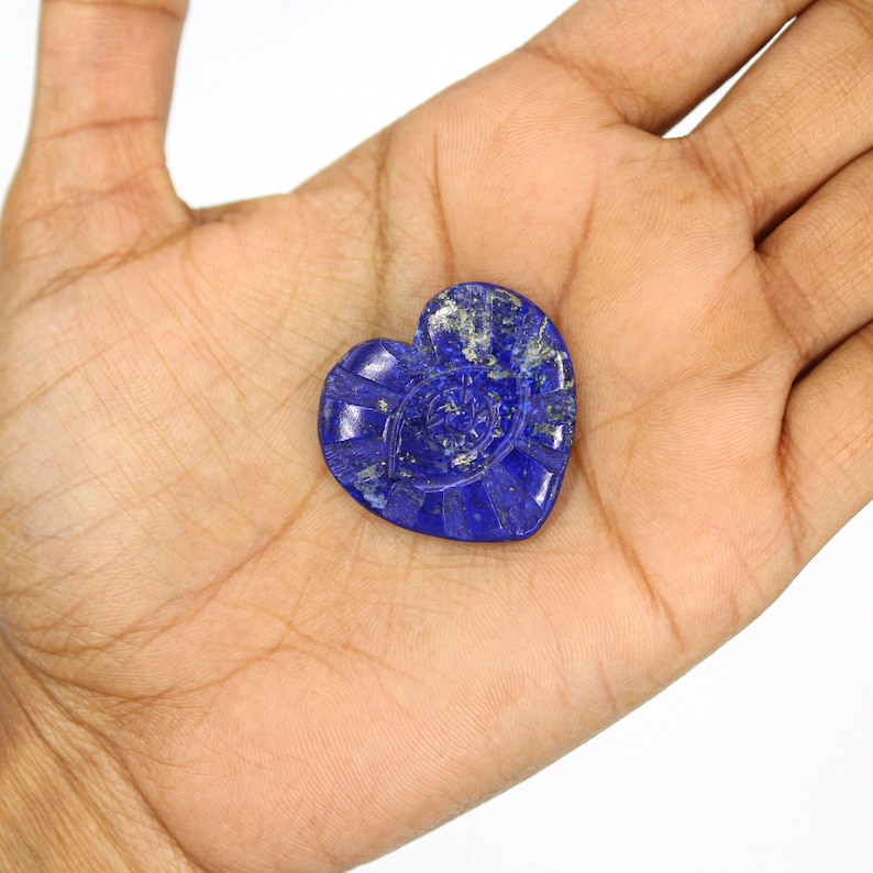 Puede incluir: Una piedra de lapisl&aacute;zuli azul oscuro en forma de coraz&oacute;n, con detalles tallados. La piedra se sostiene en una mano, resaltando su intrincado dise&ntilde;o y color. El dise&ntilde;o tallado incluye l&iacute;neas radiales y un motivo circular central.