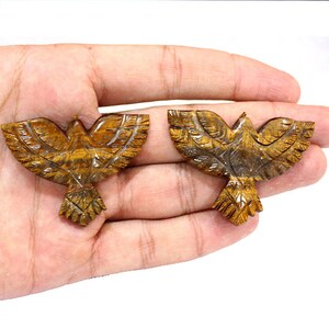 May include: Two carved stone eagle figurines with outstretched wings. The eagles are a warm brown color with detailed feather carvings. The figurines are held in a hand against a white background.
