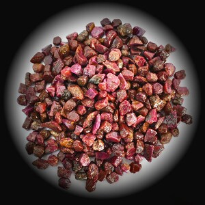 May include: A pile of rough-cut ruby gemstones. The stones vary in size and shape, displaying a range of red hues, from deep crimson to lighter shades. Some stones have a translucent quality, while others appear more opaque.