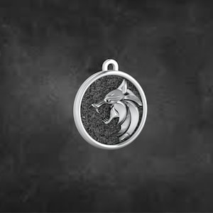 May include: A silver pendant featuring a detailed relief of a stylized animal head, possibly a wolf or similar creature, with an open mouth and sharp teeth. The pendant has a textured, dark grey background within a circular frame and a loop for a chain.
