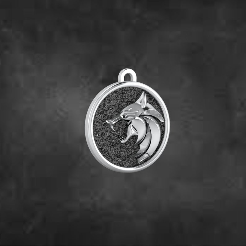 May include: A silver pendant featuring a detailed relief of a stylized animal head, possibly a wolf or similar creature, with an open mouth and sharp teeth. The pendant has a textured, dark grey background within a circular frame and a loop for a chain.