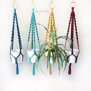 May include: Three macrame plant hangers in teal, yellow, and burgundy with white pots. The plant hangers are made with knotted cords and have tassels at the bottom. The plant hangers are hanging from the ceiling.