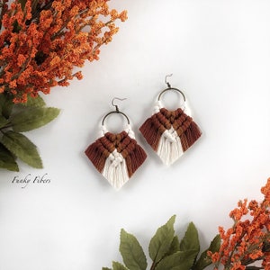 May include: A pair of handmade macrame earrings with a geometric design. The earrings are made with white and brown cord and have a fringe detail. The earrings are hanging from gold hoops.