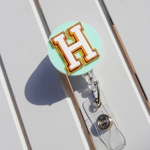 May include: A badge reel with a mint green circular base. The reel features a white letter "H" with a gold outline. The reel has a clear plastic strap and a metal clip for attaching to clothing.