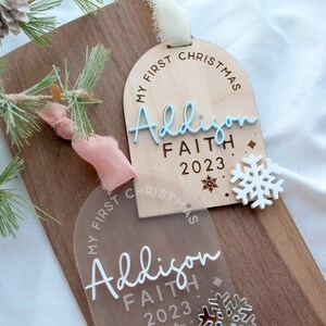 May include: Two Christmas ornaments with the text "My First Christmas" and the name "Addison Faith 2023". One ornament is clear acrylic, the other is wood. A white snowflake is next to the wooden ornament. A sprig of pine and ribbon are in the background.