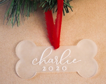 Puede incluir: Adorno en forma de hueso con el nombre "Charlie" y el año "2020" en escritura blanca. El adorno cuelga de una cinta roja, sobre un fondo marrón con agujas de pino verdes.