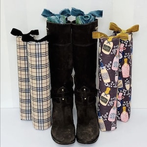 May include: A pair of brown suede boots with a black buckle detail. The boots are being stored with two pairs of boot shapers. One pair is plaid and the other pair has a champagne bottle print.