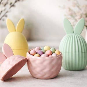 Puede incluir: Contenedores decorativos con forma de conejos de Pascua. Los contenedores son de colores pastel: rosa, amarillo y verde. El contenedor rosa está abierto y lleno de caramelos moteados de colores. El fondo incluye jarrones blancos y ramas.