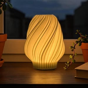 Op de afbeelding: Een crèmekleurige, gedraaide keramische lamp staat op een houten oppervlak bij een raam. De lamp heeft een gestructureerd, geribbeld ontwerp en straalt een zachte, warme gloed uit. Potplanten zijn aan weerszijden van de lamp zichtbaar.
