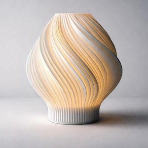 May include: A white, sculptural lamp with a unique design. The lamp features a series of curved, layered elements that create a swirling pattern. The interior glows with a warm light, and the base has vertical ridges.