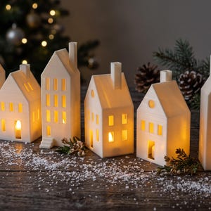 May include: A set of six white ceramic house-shaped tea light holders with cut-out windows and chimneys. Each structure features a warm, glowing yellow light emanating from within, creating a soft illumination through the rectangular openings.