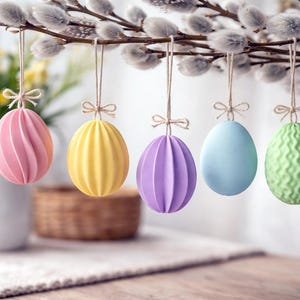 Può includere: Cinque decorazioni pasquali a forma di uovo dai colori pastello appese a un ramo di salice. Le uova sono rosa, gialle, viola, blu e verdi, ognuna con un design strutturato unico. Un boccale bianco e un cestino sono sullo sfondo.