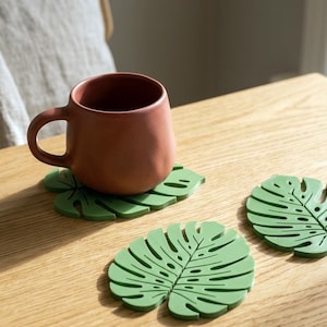 May include: A terracotta-colored mug rests on a green, leaf-shaped coaster. Two additional green leaf coasters are arranged nearby on a light-colored wooden surface. The coasters have a detailed vein pattern.