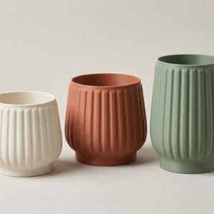 May include: Three cylindrical planters with vertical ribbed textures. The left planter is off-white, the center planter is terracotta orange, and the right planter is sage green. Each vessel features a smooth, matte finish and a wide, open top.