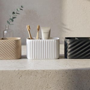 May include: Three modern toothbrush holders in beige, white, and black, each with a unique textured design. The beige holder has a diagonal wave pattern, the white has vertical lines, and the black has a diagonal wave pattern. Each holds toothbrushes.