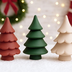 May include: Three decorative Christmas tree figurines in shades of red, green, and cream. The trees have a layered design and are set against a backdrop of a wreath, Christmas stocking, and string lights. The trees are approximately 10 cm tall.