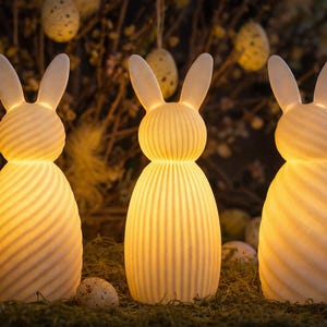Op de afbeelding: Drie verlichte keramische konijnenbeeldjes met lange oren en getextureerde lichamen. De konijnen zijn van binnenuit verlicht en geven een warme gloed. De scène bevat decoratieve paaseieren en bladeren, wat een feestelijke paasweergave creëert.