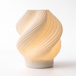 May include: A cream-colored, sculptural table lamp with a unique, twisted design. The lamp features a ribbed texture and emits a warm, inviting glow. The base is a solid, circular shape. A modern home decor accent.