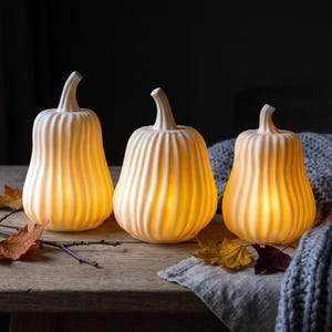 Op de afbeelding: Drie gebroken witte, geribbelde, pompoenvormige lampen met een matte afwerking. Elke pompoen heeft een gebogen steel aan de bovenkant en straalt een warme, zachte gele gloed uit. De pompoenen staan op een rij op een houten oppervlak met herfstbladeren.
