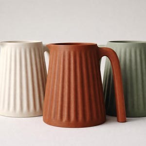 May include: Three ceramic pitchers in cream, terracotta, and sage green. Each pitcher has a ribbed design and a large handle. The pitchers are displayed on a white surface, showcasing their minimalist design and neutral color palette.