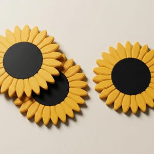 May include: Yellow sunflower-shaped coasters with black centers. The coasters are arranged on a light beige surface. The petals are a bright yellow, and the centers are a solid black. The coasters are a decorative item.