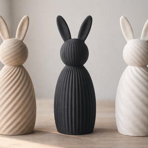 Könnte beinhalten: Drei dekorative Hasenfiguren in Beige, Schwarz und Weiß. Jeder Hase hat einen runden Kopf und einen Körper mit einem Spiralmuster. Die Hasenohren sind aufrecht. Auf einer Holzoberfläche platziert.