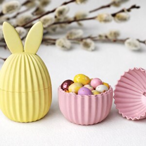 Peut inclure: Un récipient jaune en forme d'œuf avec des oreilles de lapin, un bol rose rempli de bonbons colorés et un couvercle rose à bord festonné. Les articles sont disposés sur une surface blanche avec un arrière-plan flou de branches.