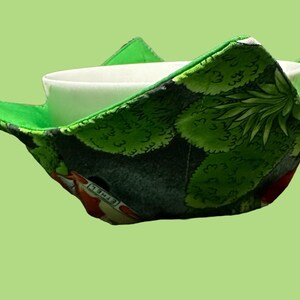 May include: A green and black fabric bowl cosy with a leafy pattern. The cosy is holding a white ceramic bowl.